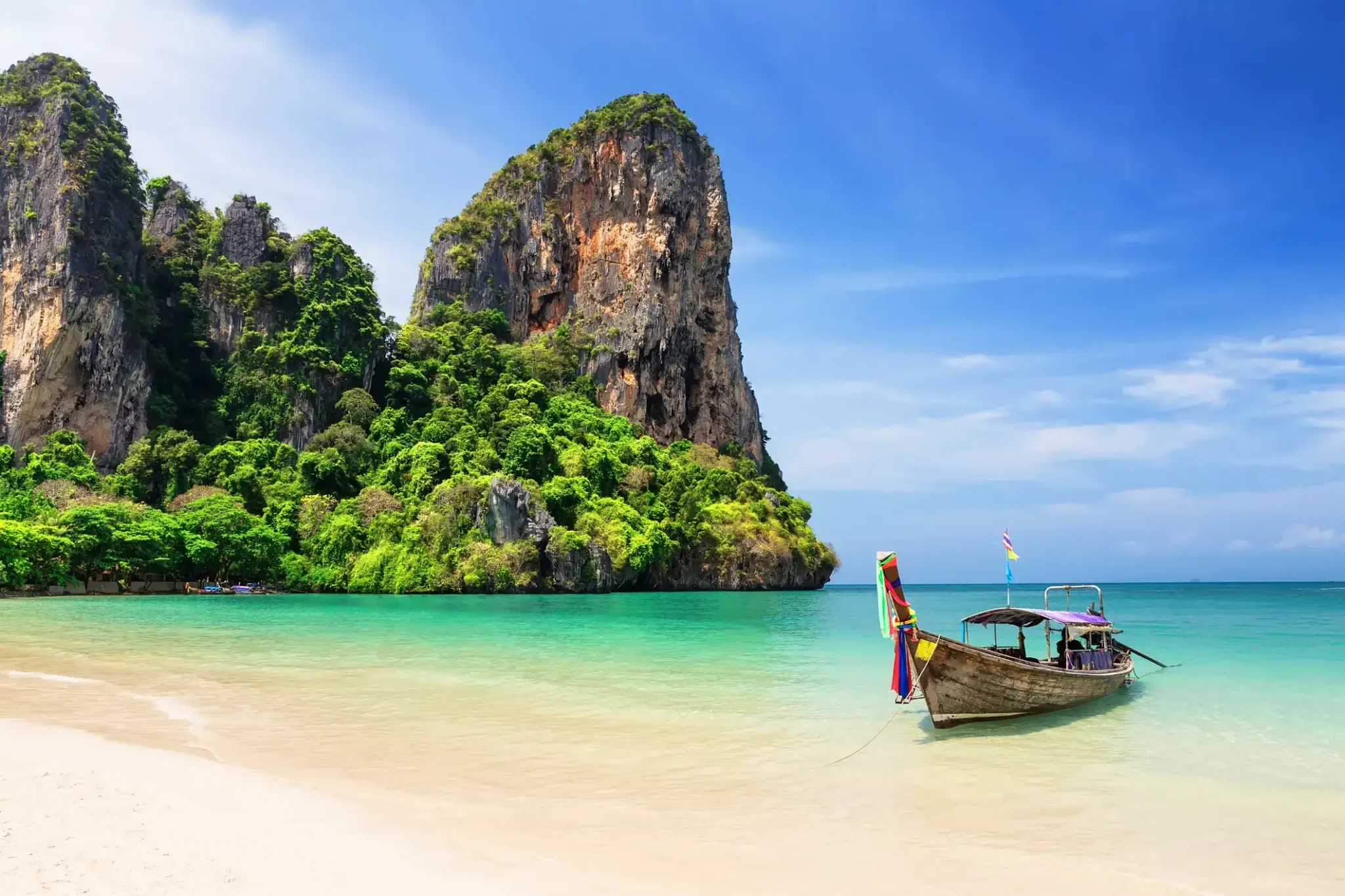 A traditional wooden boat anchored on a tropical beach with turquoise waters and towering limestone cliffs in Thailand.