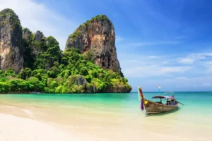 A traditional wooden boat anchored on a tropical beach with turquoise waters and towering limestone cliffs in Thailand.