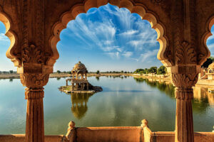 A scenic view of Gadisar Lake in Jaisalmer, Rajasthan, framed by ornate sandstone arches, with a chhatri pavilion reflecting in the calm water under a blue sky.