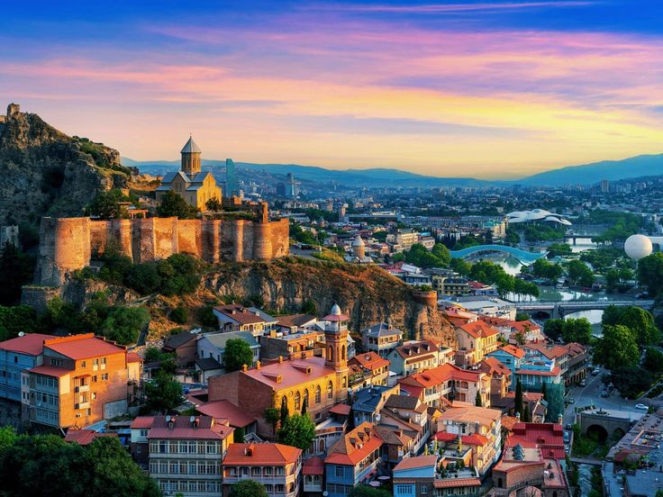 A panoramic view of Tbilisi, Georgia, showing colorful rooftops, historic buildings, and the ancient Narikala Fortress overlooking the city under a clear blue sky.