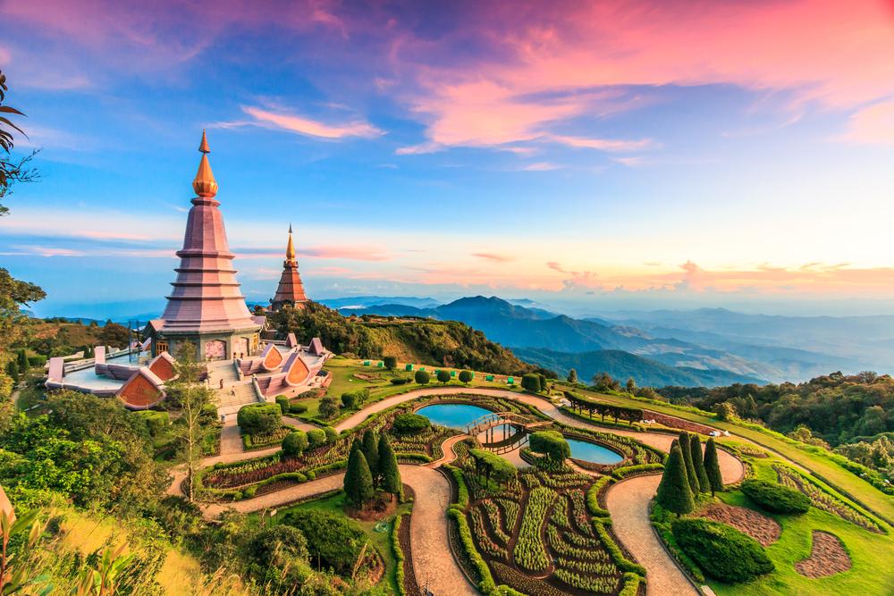 A scenic view of twin pagodas atop a hill surrounded by beautifully landscaped gardens, pathways, and ponds, with distant mountain ranges under a vibrant pink and blue sunset sky.
